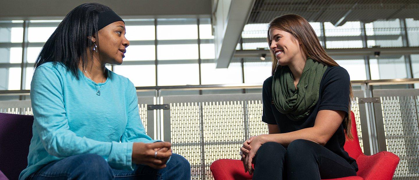 Two students seated in chairs, talking to each other.